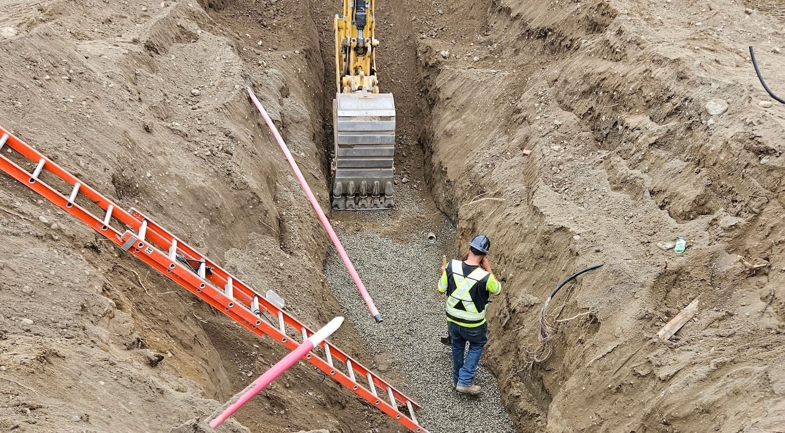 Excavator places bedding aggregate while civil laborer grades trench base in preparation for underground service installation.