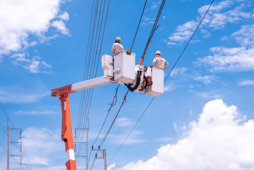 Two electricians on bucket boom truck are connecting electrical cable lines against blue sky background.